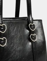 Cartera tipo city de cuerina color negro con efecto charol. Tiene manijas superiores con apliques metálicos en forma de corazón, cierre superior y bolsillos laterales con trenzado decorativo.