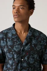 Camisa de hombre de manga corta con estampado floral en tonos azules sobre fondo negro. Tiene cuello tipo cubano, cierre frontal con botones y un pequeño logo rojo bordado en el pecho.
