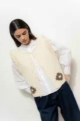 Chaleco tejido color crema con cuello redondo y borde con flecos. Presenta apliques de flores marrones en la parte inferior.