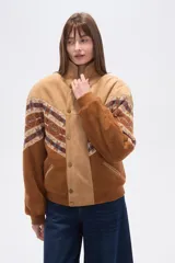 Chaqueta holgada de poliéster en color tostado con forro de corderito, cuello de solapa y cierre frontal con botones. Presenta mangas con apliques de diseño geométrico en tonos marrones y azules.
