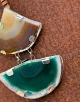 Collar con colgante de dos piedras naturales irregulares engarzadas en metal plateado y dorado, suspendidas de un cordón de cuero marrón con detalle de nudo corredizo. Las piedras tienen vetas en tonos naranja, marrón y blanco.