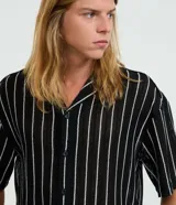 Camisa masculina modelo boxy, confeccionada en punto, con cuello polo, manga corta, cierre por botones y estampa de rayas verticales blancas sobre fondo negro.