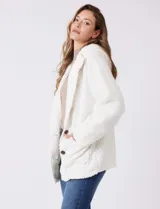 Campera tipo teddy de textura suave y esponjosa en color blanco, con cierre frontal mediante botones y detalles de apliques en cuero sintético negro en la solapa. Presenta bolsillos frontales y un diseño cómodo y abrigado.