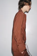Camisa marrón de Zara Woman Collection confeccionada en hilatura con mezcla de lyocell. Tiene cuello redondo, manga larga acabada en puño, bajo asimétrico con aberturas laterales y cierre frontal con botones.