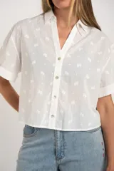 Camisa blanca corta de algodón con bordado de flores, cuello clásico, cierre frontal con botones y mangas cortas con dobladillo.