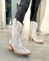 Bota texana de caña alta color marrón claro, confeccionada en cuero vacuno con detalles de bordado y apliques en relieve. Tiene cierre lateral, taco de madera con folia de cuero y suela de goma. La horma es de punta fina, ancha y cómoda.