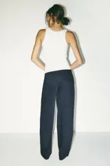Pantalón negro con finas rayas verticales blancas, de tiro medio y corte ancho. Presenta bolsillos delanteros y traseros, cintura doble tipo bóxer con botones y cierre frontal con cremallera y ganchos metálicos. Detalle de franja marrón en los laterales.