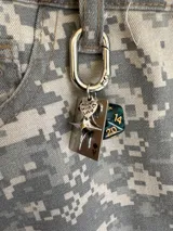 Llavero metálico plateado con mosquetón, que incluye varios dijes: una placa rectangular con el As de Picas, un dado poliédrico verde oscuro y dorado, una estrella de cuatro puntas, y un dije pequeño con forma de corazón que dice "made with love".