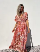 Vestido largo de corte evasé con estampado floral en tonos rosados y anaranjados. Tiene escote en V cruzado, mangas cortas con volantes y falda con volantes asimétricos.