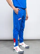 Pantalón de jogging azul marino con puños ajustados, cintura elástica con cordón y bolsillos laterales. Presenta una franja blanca con detalles en rojo a lo largo de la pierna, el escudo del Club Nacional de Football y el logo de Umbro.