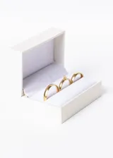 Set de tres anillos dorados, dos alianzas lisas y un cintillo con una piedra engarzada, presentados en un estuche blanco.
