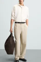 Pantalón de corte holgado (wide fit) en color beige, confeccionado en tejido con mezcla de lyocell. Presenta pliegues frontales en la cintura, bolsillos laterales y bolsillos traseros de doble vivo. Cuenta con cierre frontal de cremallera y botón.