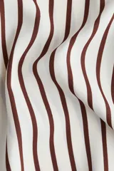 Camisa de crepé color crema con rayas verticales marrones, corte oversized, cuello clásico, cierre frontal con botones ocultos, mangas largas con puños abotonados, pliegue de caja invertido en la espalda y bajo recto.