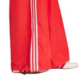 Pantalón Adidas rojo de corte ancho con cintura elastizada y cordón ajustable. Presenta las tres tiras blancas características de la marca en los laterales y el logo bordado en la pierna izquierda.