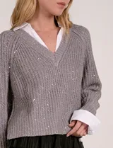 Buzo de tejido de punto gris, con cuello en V y detalles de lentejuelas. Se lleva superpuesto a una camisa blanca con cuello y puños visibles.