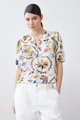 Camisa blanca con estampado floral en tonos azul, amarillo, rosa y marrón. Tiene cuello mao, mangas cortas abullonadas y cierre frontal con botones.