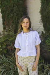 Camisa blanca de lino con alforzas verticales, cuello fruncido y mangas abullonadas con elástico. Los botones están bordados a mano con hilo de algodón. Tiene corte recto con pequeño volado en la base.