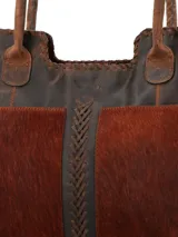 Bolso tote de cuero marrón oscuro con paneles frontales de pelo (pelo vacuno) en tono marrón rojizo. Presenta costuras decorativas en los bordes y un trenzado central de cuero oscuro. Tiene asas de hombro de cuero desgastado.