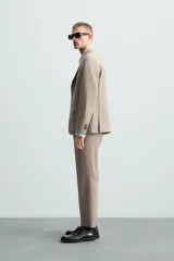 Pantalón de vestir para hombre, color beige claro con textura de tejido de lana. Presenta un corte regular fit, con pliegues marcados en el frente, bolsillos laterales y bolsillos traseros de doble vivo con botón.