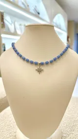 Gargantilla con cuentas cilíndricas de color azul y cuentas transparentes más pequeñas. Dije colgante en forma de estrella de ocho puntas con incrustaciones de circonitas.