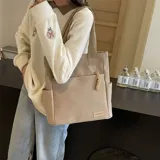 Cartera tipo tote de lona en color beige con correas de cinta marrón. Presenta un compartimento principal con cierre, un bolsillo frontal abierto y bolsillos laterales para mayor organización.