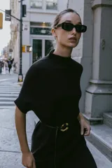 Vestido negro de tejido de punto, corte midi y silueta holgada, con cuello alto acanalado y mangas cortas tipo murciélago. Se ajusta a la cintura con un cinturón fino de efecto piel negro con hebilla dorada en forma de C.