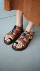 Sandalias de cuero color marrón con tiras cruzadas y detalles en negro, con hebillas doradas y suela plana.