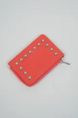 Billetera pequeña con cierre de cremallera, color coral o naranja vibrante, decorada con tachas metálicas plateadas en forma de media esfera dispuestas en un patrón curvo en la parte frontal.