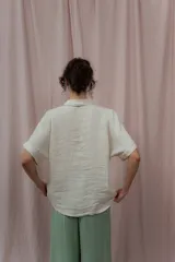 Camisa color crudo confeccionada en lino, de corte holgado, con cuello clásico, mangas cortas con dobladillo y cierre frontal con botones. Presenta detalle de pliegues verticales en la parte frontal.