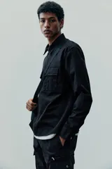 Sobrecamisa negra de algodón con cuello inglés, cierre bidireccional, bolsillos superiores con solapa y botones a presión, canesú en la espalda, bajo recto con aberturas laterales y mangas largas con tapeta de manga y botón a presión en los puños.
