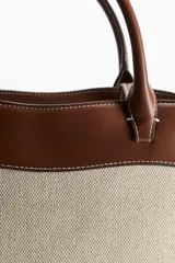 Bolso shopper de lona color beige con detalles en cuero marrón. Tiene doble asa de mano y compartimento interno con cierre.
