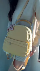 Mochila color mostaza de símil cuero con bolsillo frontal con cierre y broche, apertura superior con cierres metálicos, bolsillos laterales y asa de mano. Incluye bolsillos internos y correas regulables para colgar en la espalda.