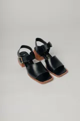 Sandalias negras de cuero con diseño minimalista y taco ancho geométrico en tono madera. La capellada lisa aporta una estética depurada y moderna, mientras que la pulsera con hebilla metálica ajustable asegura un calce firme y confortable.