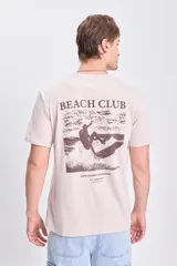 Remera de manga corta color beige con estampa en la espalda con la frase "Beach Club" y un diseño de un surfista en la playa.