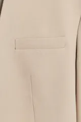 Blazer color beige, sin solapa, de manga larga con bolsillos de vivo en el delantero y cierre frontal con gancho metálico.