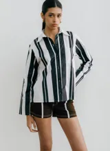 Camisa blanca con rayas verticales negras, de manga larga y corte recto. Tiene cuello clásico, cierre con botones y bolsillo en el pecho.