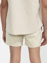 Short de baño color beige con una franja vertical marrón en el lateral. Tiene cintura elástica con cordón ajustable, bolsillos laterales y trasero con cierre, y suspensor de malla fina hipoalergénica.