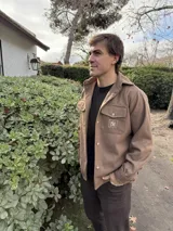 Camisa marrón de lana con corte clásico, cuello camisero, cierre frontal con botones, dos bolsillos de parche con solapa abotonada en el pecho y puños abotonados. Presenta un pequeño logo bordado en uno de los bolsillos.