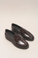 Mocasín negro de cuero vacuno con acabado charol, suela de goma y forro y plantilla de cuero vacuno.