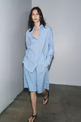 Camisa celeste con rayas verticales blancas, de corte amplio, cuello clásico, manga larga con puños y cierre frontal con botones.