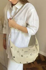 Cartera tipo bandolera de cuero sintético color celeste, con forma de media luna y tachas metálicas redondas plateadas en el frente. Posee correa ajustable y placa metálica con logo en la parte superior.