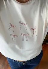 Remera blanca de algodón con cuello redondo y mangas largas. Estampado frontal con diseño de moños rojos.