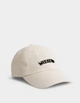 Gorra de gabardina color crudo con visera curva y detalle de roturas. Bordado frontal con la palabra "Weekend" en negro.