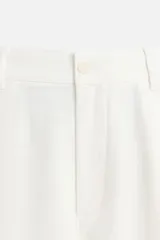 Pantalón blanco de corte relajado con pliegues frontales, bolsillos laterales y bolsillos traseros de vivo. Cierre frontal con cremallera y botón.