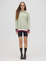 Sweater de punto color verde claro con cuello polo con cierre de cremallera, mangas largas y diseño con trenzas. Puños acanalados con rayas en color borgoña y blanco.