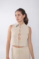 Top tejido color beige con espalda calada y cuello mao. Presenta cierre frontal con cordones y detalle de broches decorativos en el cuello.