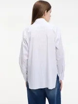 Camisa blanca a rayas verticales finas, con cuello clásico y cierre frontal con botones. Presenta apliques de pedrería en el cuello y en la tapeta de los botones.