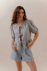 Blusa lisa color gris claro, con escote redondo y lazos frontales regulables. Presenta mangas cortas abullonadas con elástico en los puños y un leve frunce en la cintura que crea un efecto peplum.