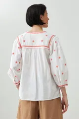 Blusa blanca de corte holgado con bordados de estrellas y detalles geométricos en color coral.