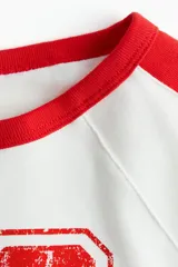 Remera corta blanca de punto acanalado fino de algodón con mangas raglán y ribetes en rojo. Estampado frontal con el número 28 en rojo.